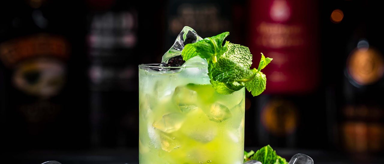 Green Negroni cocktail drink with gin, aperitif, liqueur, mint and ice. Black bar counter background with bottles