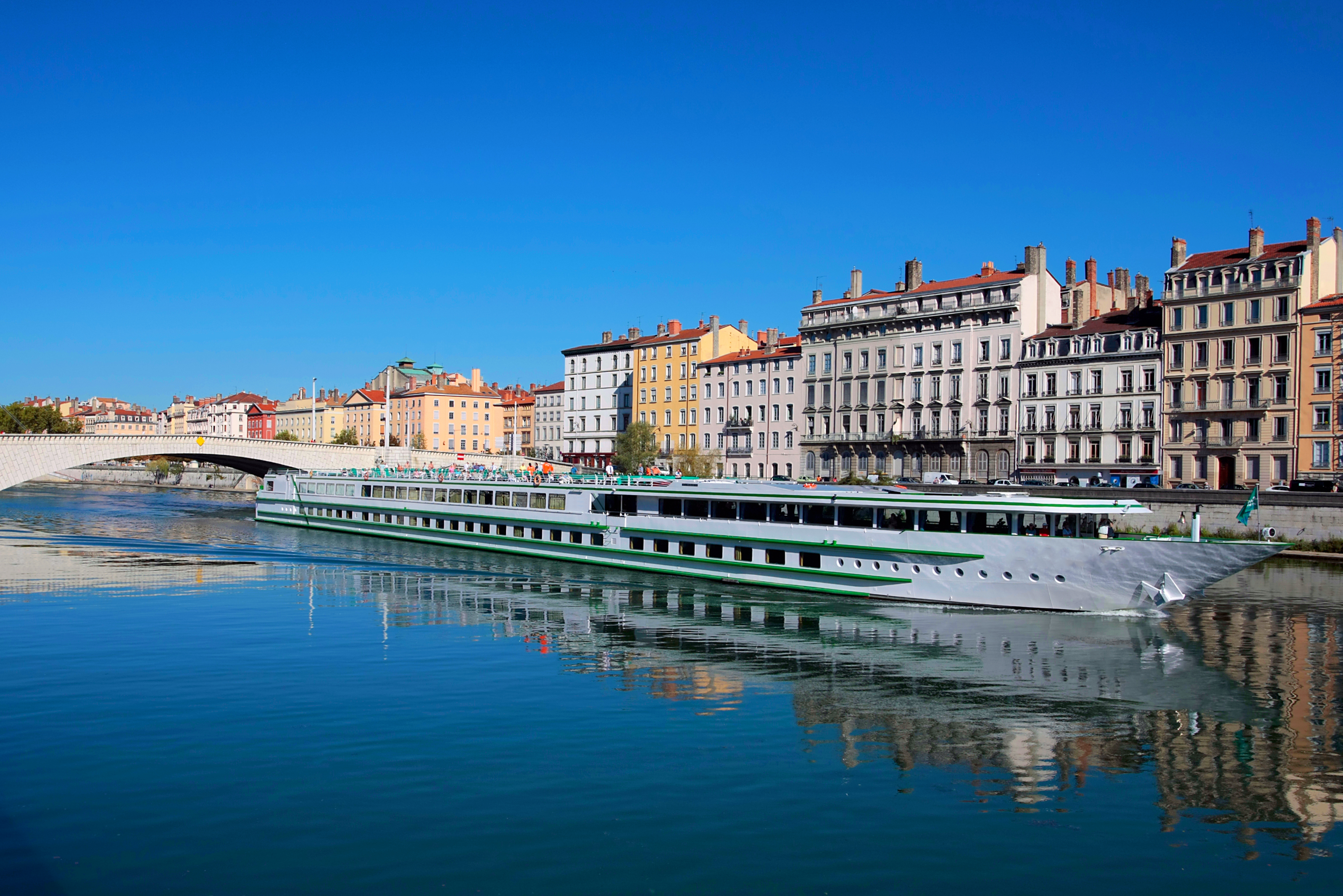 River cruise ship sailing along the Saône River in Lyon, France, with colourful historic riverside buildings, a bridge in the background, and clear blue skies reflected in the calm water.