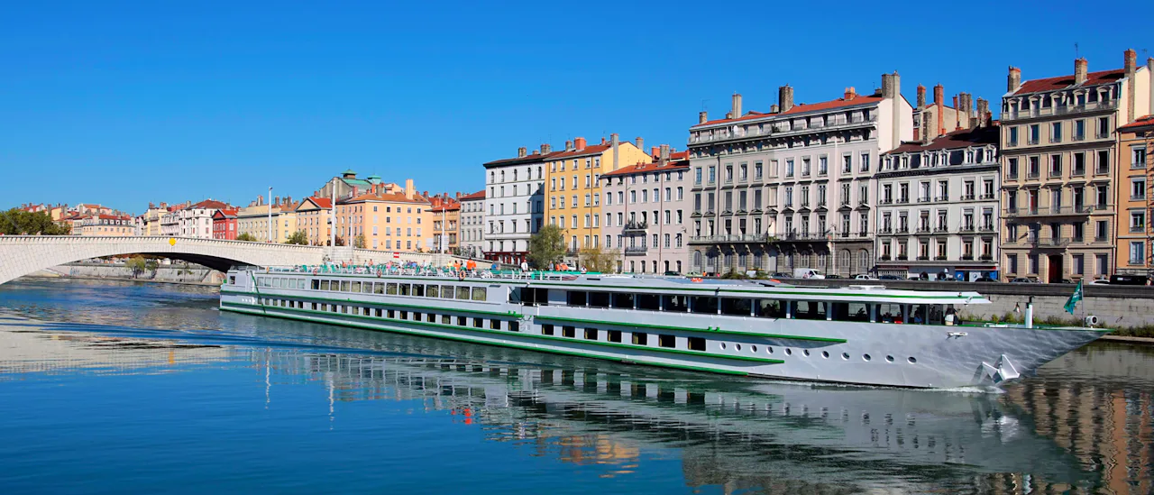 River cruise ship sailing along the Saône River in Lyon, France, with colourful historic riverside buildings, a bridge in the background, and clear blue skies reflected in the calm water.