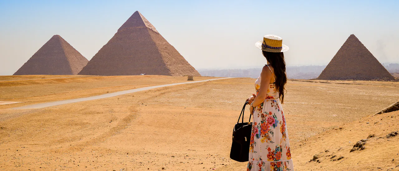 Woman in a floral dress and sun hat standing in the desert, looking towards the Pyramids of Giza in Egypt under a clear blue sky.